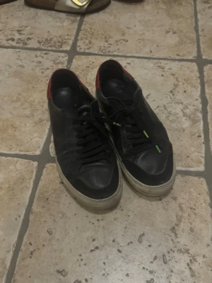 Svarta låga Arigatos med röd detalj - Snygga svarta sneakers med lågt skaft och röd detalj på hälen. Skorna har svarta snören med gröna spetsar och en vit sula som ger en klassisk look. Perfekta för dig som gillar enkel men stilren design. Materialet ser ut att vara läder eller konstläder.