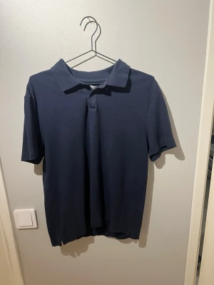 Mörkblå pikétröja från These Glory Days - En klassisk mörkblå pikétröja frånThese Glory Days med krage och korta ärmar. Tillverkad i mjuk bomull som känns skön mot huden. Perfekt till jeans eller shorts för en avslappnad look nu till sommaren.