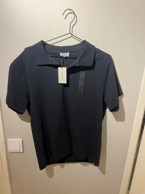 Mörkblå pikétröja från Sandö med prislapp kvar - Klassisk mörkblå pikétröja från Sandö med krage och korta ärmar. Tröjan har en stilren look utan synliga loggor på bröstet och är tillverkad i mjukt bomullsmaterial. Perfekt för en clean och enkel stil. Säljes på grund av fel storlek, nypris 499, mitt pris 299