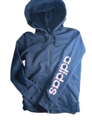 Mörkblå hoodie från Adidas - Snygg mörkblå hoodie från Adidas med dragkedja framtill och stor vit Adidas-logga längs ena ärmen. Tröjan har huva med dragsko och två fickor fram. Tillverkad i mjukt bomullsmaterial som är skönt att ha på sig. Perfekt för en avslappnad och sportig stil.