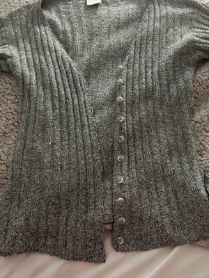 Grå ribbad kofta med knappar - Säljer en grå ribbad kofta med v-ringning och vita knappar framtill. Koftan har ett mjukt, stickat material och långa ärmar. Perfekt för lager på lager och enkel att matcha med olika outfits. Storlek 34/36 men skulle säga att det är som small 