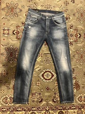Blå slim fit jeans från Dondup - Säljer ett par blå slim fit jeans från Dondup med snyggt slitna detaljer och klassisk femficksmodell. Jeansen har en tvättad look med ljusare partier och är tillverkade i ett mjukt denimtyg. Perfekta för en avslappnad och trendig stil.