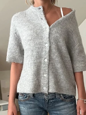 Ljusgrå kofta från Gina Tricot - Mysig ljusgrå kofta från Gina Tricot i ribbstickad design. Koftan har korta ärmar, rund halsringning och knäpps med vita knappar framtill. Tillverkad i ett mjukt, fluffigt material som känns lyxigt och skönt mot huden.