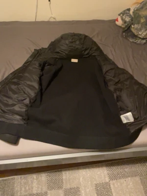 Svart dunväst från Moncler - Snygg svart dunväst från Moncler med klassisk quiltad design och diskret logga på insidan. Västen har en hög krage och är fodrad med dun för extra värme. Perfekt att slänga över en hoodie eller tröja när det är kyligt ute.