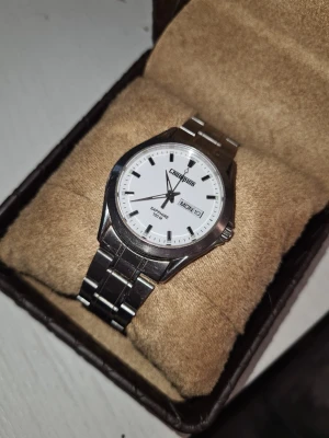 Silverfärgad Champion klocka - Stilren klocka från Champion med vit urtavla och dag- och datumvisning. Armbandet är i blankt silverfärgat metall och har ett klassiskt spänne. Urtavlan är enkel med svarta markeringar och visare. Perfekt accessoar för dig som gillar en clean look.