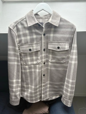 Beige-rutig overshirt med fickor - Stilren overshirt i beige och grått rutmönster. Skjortjackan har två bröstfickor med lock och svarta knappar framtill. Klassisk krage och långa ärmar med knapp vid ärmslut. 