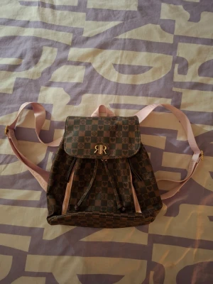 Miniryggsäck river island - Miniryggsäck från river island med färgerna brun och ljusrosa. Jättefin och knappt använd! Köpt ny för ca 400-500kr, och är i mycket fint skick. Axelbanden går att justera genom att förminska och förstora. Hör av dig vid funderingar.