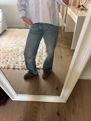 Blå slitna bootcut jeans - Säljer ett par blå bootcut jeans med slitningar och hål på både fram- och baksida. Jeansen har en avslappnad passform, klassisk femficksdesign och är gjorda i denim. Perfekta för en trendig och avslappnad look.