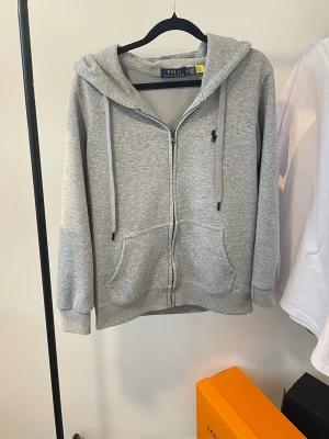 Grå Polo Ralph Lauren Zip-Hoodie - Säljer nu en klassisk grå zip-hoodie från Polo Ralph Lauren. Tröjan har den ikoniska svarta hästen broderad på bröstet och en mjuk, skön insida. Perfekt passform som funkar till både jeans och mjukisbyxor. 