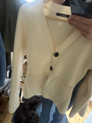 Krämvit kofta från Gina Tricot - Mysig krämvit kofta från Gina Tricot med djup V-ringning och tre stora svarta knappar framtill. Koftan har två framfickor och är stickad i ett mjukt material. Perfekt att slänga över en t-shirt eller linne för en chill look.