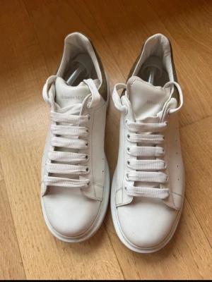 Vita Alexander McQueen sneakers - Stilrena vita sneakers från Alexander McQueen med chunky sula och breda vita skosnören. Skorna har diskreta hål på sidorna och en kontrasterande grön detalj på hälen. Tillverkade i läder för en lyxig känsla och look. Storlek 42 typ 43. Pris kan diskuteras. Hör av dig!