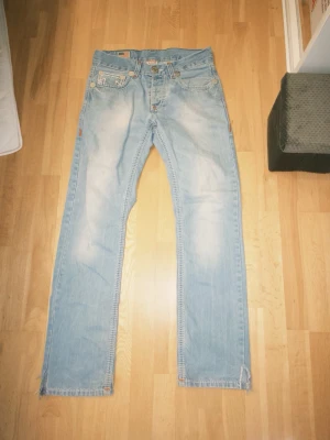 Ljusblå True Religion jeans - Säljer ett par ljusblå jeans från True Religion med raka ben och klassiska kontrastsömmar. Jeansen har fem fickor, tydliga broderier på bakfickorna och läderpatch i midjan. Tillverkade i bomull med slitna detaljer för en cool look.