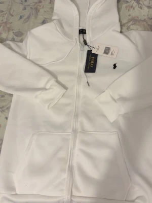 Vit hoodie från Polo Ralph Lauren - Snygg vit hoodie från Polo Ralph Lauren med dragkedja framtill och klassisk huva. Har två stora fickor fram och den ikoniska lilla svarta loggan broderad på bröstet. Perfekt för en clean och stilren look.