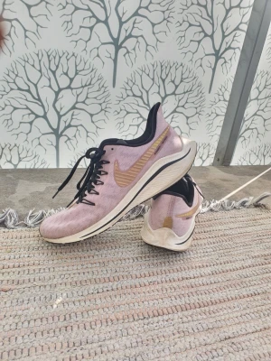 Nike rosa sneakers med guld swoosh - Snygga rosa Nike sneakers med guld swoosh på sidan och svarta detaljer. Skorna har svarta snören och en vit sula med svart kant. Perfekta för dig som vill ha en sportig och trendig look.