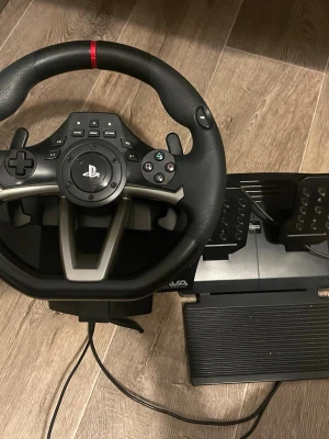 Ratt och pedaler till PlayStation - Säljer en svart och silverfärgad ratt med pedaler från Hori, designad för PlayStation. Ratten har gummiklädda handtag, PlayStation-knappar och växelpaddlar. Perfekt för racing- och bilspel, kopplas in med kabel.