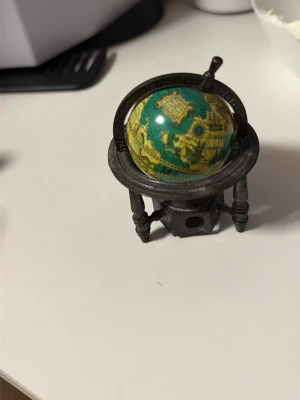 Miniatyrglob i antik stil - En liten dekorativ jordglob i antik stil med gröna och gula färger. Globens stativ är i mörkgrått metallutseende med tre ben och detaljerad ram runt globen. Perfekt som prydnad eller samlarobjekt till skrivbordet eller hyllan.