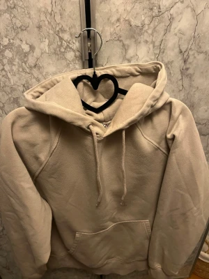 Beige hoodie med ficka framtill - En stilren beige hoodie med huva och dragsko, samt en stor känguruficka framtill. Hoodien har raglanärmar och är tillverkad i mjukt material, perfekt för en avslappnad look. Passar dig som gillar enkel och clean stil.