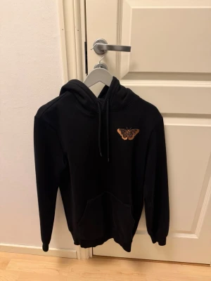 Svart hoodie med fjärilstryck - Svart hoodie med huva och dragsko, samt en snygg orange fjäril på bröstet. Klassisk känguruficka framtill och mjukt material som känns skönt mot huden. Perfekt för dig som gillar stilrena plagg med en unik detalj.