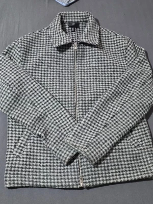 Gråmelerad jacka från H&M - Snygg gråmelerad jacka från H&M med klassiskt hundtandsmönster. Jackan har dragkedja framtill, två sidofickor och en rak passform. Perfekt för dig som gillar stilrena och tidlösa plagg.