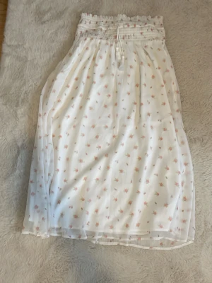 Vit blommig långkjol från Gina Tricot - Söt vit långkjol från Gina Tricot med små rosa och gröna blommor. Kjolen har smock i midjan med knytband och är tillverkad i ett lätt och luftigt material, perfekt för varmare dagar.