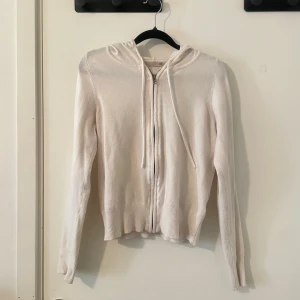 Vit kofta - Säljer en stilren vit kofta med dragkedja framtill och justerbar huva med snören. Hoodien har långa ärmar och ribbade muddar vid ärmslut och nederkant. Perfekt för en avslappnad look och enkel att matcha med olika outfits.