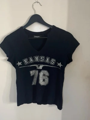 Mörkblå Kansas 76 t-shirt med v-ringning - Cool mörkblå t-shirt från Atmosphere med v-ringning och tryck i vitt och silver med texten 'KANSAS 76' och stjärnor. Kort ärm och något croppad passform. Perfekt för en avslappnad och sportig stil. 