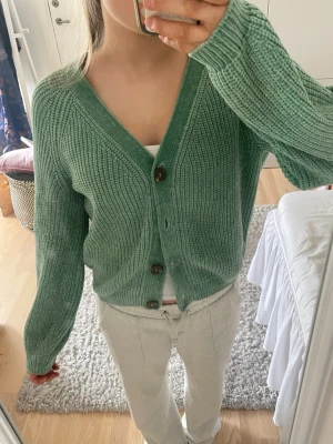 Grön stickad kofta med knappar - Mysig grön stickad kofta med v-ringning och stora bruna knappar framtill. Koftan har lång ärm och en avslappnad passform, perfekt att slänga på sig över en t-shirt. Materialet är grovstickat och känns mjukt mot huden. Sitter som en xs