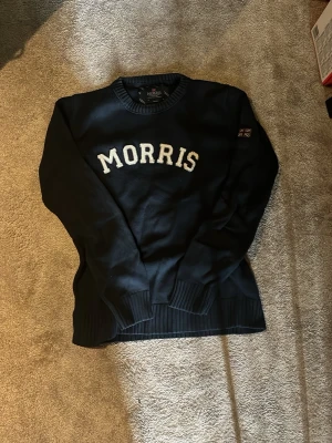 Svart Morris tröja med tryck - Snygg svart tröja från Morris med vit broderad text på bröstet och en liten brittisk flagga på ärmen. Tröjan har rund halsringning och ribbade muddar vid ärmslut och nederkant. Perfekt för en stilren och klassisk look.