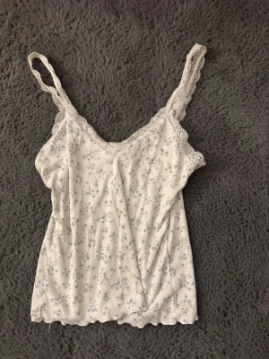 Vit blommig spetsbody - Söt vit body med små blå blommor och tunna axelband. Spetsdetaljer längs urringningen och axelbanden ger en romantisk känsla. Tillverkad i mjukt, ribbat tyg som känns bekvämt mot huden. Perfekt för dig som gillar en feminin och enkel stil.