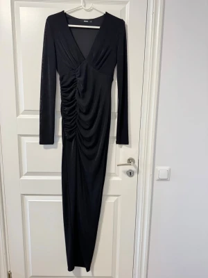 Svart långklänning med drapering - Elegant svart långklänning från NA-KD med v-ringning, långa ärmar och snygg drapering längs sidan. Kläningen har en låg slits. Och  har en figurnära passform.