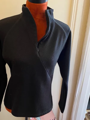 Svart halvzip tröja i funktionsmaterial 40 - Svart Craft tröja med långa ärmar och hög krage. Tröjan har en dragkedja framtill som går ner en bit på bröstet. Kragen är designad för att ge extra värme och skydd. Tröjan har en strukturerad yta. 100% polyester Längd: 56 cm Bystvidd: 48 cm  Inga bilder på mig! Katter finns i hemmet