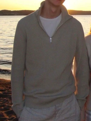 Beige ribbad halvzip-tröja - Säljer en stilren beige ribbad tröja med halvzip och hög krage. Tröjan har långa ärmar och är tillverkad i ett mjukt stickat material som ger en avslappnad men ändå snygg look. Perfekt att bära över en t-shirt för en clean och enkel stil.