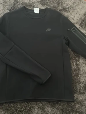 Nike Tech Tröja - Svart sweatshirt från Nike med diskret logga på bröstet och en praktisk ficka med dragkedja på ena ärmen. Tröjan har rund halsringning och långa ärmar, perfekt för en sportig och clean look.