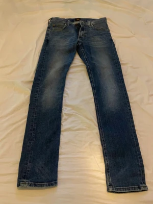 Blå slim fit jeans från Lee - Snygga blå slim fit jeans från Lee med klassisk femficksdesign och diskreta slitningar på låren. Jeansen har normal midja och Lee-logga på knapp och etikett. Perfekta för en avslappnad och trendig look.
