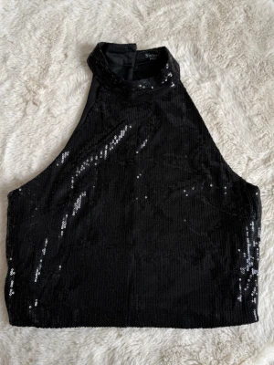 Svart paljettopp från Sisters Point XS - Snygg svart croppad topp från Sisters Point med paljetter över hela ytan. Toppen har hög hals och är ärmlös, perfekt för att sticka ut. Materialet är glittrigt och ger en cool look.