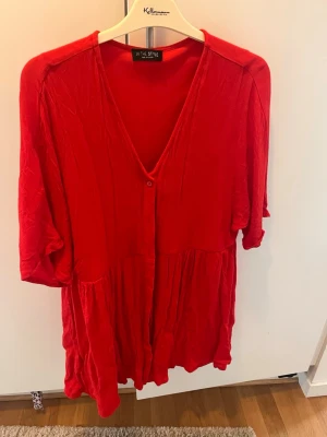 Röd klänning från In The Style - Röd klänning med v-ringning och knappar framtill från In The Style. Modellen är lös med trekvartsärm och har en avslappnad passform. Perfekt för dig som gillar färgstarka plagg med enkel design.