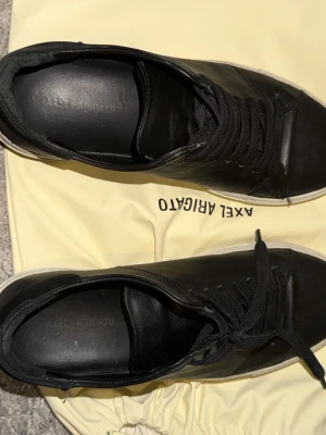 Svarta sneakers från Axel Arigato -  svarta sneakers från Axel Arigato i läder med vita sulor och svarta skosnören. Klassisk låg modell med minimalistisk design och diskret logga på sidan. Perfekta för en clean och modern look. Liten bit sliten som man ser på tredje men inget stort som man mörker till om man inte kolla nogrant 