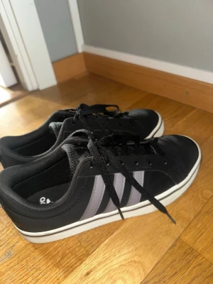 Svarta Adidas sneakers med lila ränder - Snygga svarta sneakers från Adidas med de klassiska tre vita ränderna på sidorna. Skorna har svart snörning, vit sula och är tillverkade i syntetmaterial med en stilren och tidlös design. Perfekta för en sportig och avslappnad look. Ny men använda en enda gång