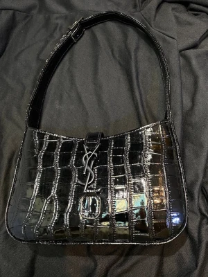 YSL väska - Snygg handväska i krokodilmönstrat lackläder Säljer nu denna eleganta och stilrena handväska i en klassisk hobo-modell. Väskan är perfekt för både vardag och fest och ger en lyxig känsla till varje outfit.