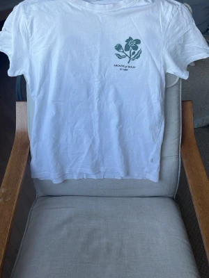 Vit t-shirt Archive Sold med blomtryck - Vit t-shirt från Archive Sold med grönt blomtryck både fram och bak. Klassisk rund hals och korta ärmar. Trycket har en cool retrostil och t-shirten är gjord i mjuk bomull, perfekt för dig som gillar stilrena och unika plagg.