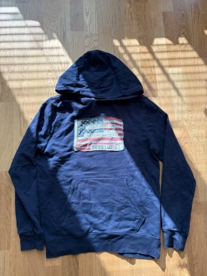 Ralph Lauren Denim supply hoodie  - Säljer en mörkblå hoodie från Ralph Lauren med stor huva och känguruficka. På bröstet finns ett stort tygmärke med amerikanska flaggan och slitna detaljer. Perfekt för dig som gillar klassisk streetstyle och coola loggor.