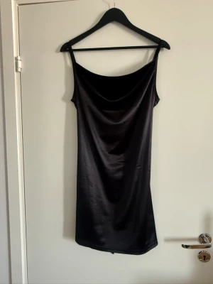 Svart satin slipklänning - Stilren svart slipklänning i satin med smala axelband och snygg draperad urringning. Klänningen har en kort längd och ett band i ryggen som kan knytas för extra passform. Perfekt för dig som gillar minimalistisk och trendig stil.