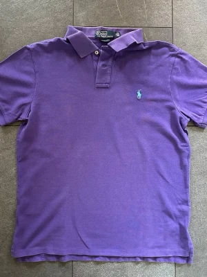 Lila pikétröja från Ralph Lauren - Snygg lila pikétröja från Polo Ralph Lauren i slim fit-modell. Tröjan har klassisk krage, två knappar och den ikoniska turkosa logotypen broderad på bröstet. Tillverkad i mjuk bomull som känns skön mot huden. Storlek L men passar som M
