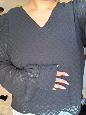 Svart transparent blus med V-ringning - Tröjan är i storlek L men passar en S/M också! Snygg svart blus med V-ringning och lång ärm. Blusen är gjord i ett transparent, mönstrat tyg med zigzagstruktur som ger en cool och trendig look. Perfekt att styla med jeans eller kjol för en modern outfit. 