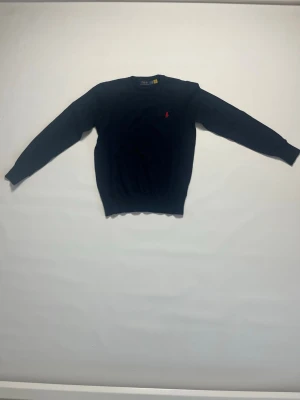 Mörkblå stickad tröja från Ralph Lauren storlek S - Mörkblå stickad tröja från Ralph Lauren med klassisk rund halsringning och röd broderad logga på bröstet. Tröjan har långa ärmar och ribbade muddar vid ärmslut och nederkant. Perfekt för en stilren och avslappnad look. Tröjan är i storlek S och den är helt ny och aldrig buren