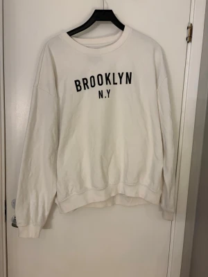 Vit sweatshirt med Brooklyn-tryck - Vit sweatshirt med svart tryck. Det finns några små fläckar på tröjan (se bilden. Tröjan är väl använd dvs det finns knottror på den.