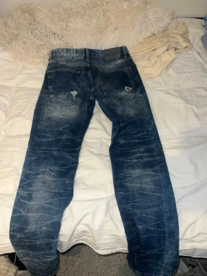 Blå slitna jeans från Red Bridge - Säljer ett par blå jeans från Red Bridge med slitningar och distressed detaljer på bakfickorna. Jeansen har en klassisk femficksmodell och är tillverkade i denim med tvättad look. Perfekta för en avslappnad och trendig stil.