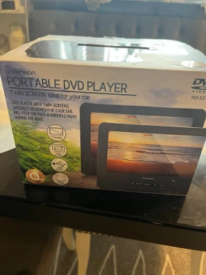 Andersson bärbar DVD-spelare med dubbla skärmar - Bärbar DVD-spelare från Andersson med två svarta skärmar, perfekt för bilen. Inkluderar fjärrkontroll och kablar. Skärmarna har en modern, rektangulär design och är tillverkade i plast. Smidig lösning för underhållning på resan.