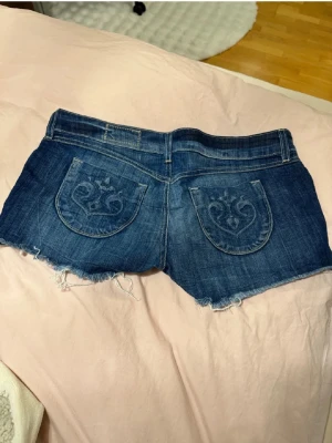 Blå jeansshorts med spetsdetaljer - Säljer ett par blå jeansshorts med råa kanter och ljusrosa spetsdetaljer framtill. Shortsen har broderade bakfickor och en klassisk femficksmodell. Perfekta för sommaren och ger en avslappnad men ändå söt look.