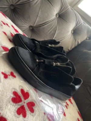 Svarta sneakers från Axel Arigato - Snygga svarta sneakers från Axel Arigato i mocka med coola silverfärgade dragkedjor på ovansidan. Skorna har en chunky svart sula och stilren design som passar perfekt till streetwear. Märkeslogga tryckt på sidan. Riktigt trendiga och unika sneakers!